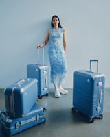 A model in a blue lace dress leans against a wall, resting her hand on a Hybrid Carry-On in Elemental Blue with more luggage of the same colour around her.