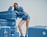 A model in a blue fleece jacket and shorts leans on a stack of Hybrid suitcases in Elemental Blue, gazing at the camera.