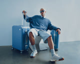 A model in a blue sweater and sunglasses lounges on a chair, surrounded by Hybrid luggage in Elemental Blue.