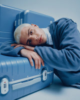 A model in a blue sweater rests their head on a Hybrid suitcase in Elemental Blue, gazing directly at the camera