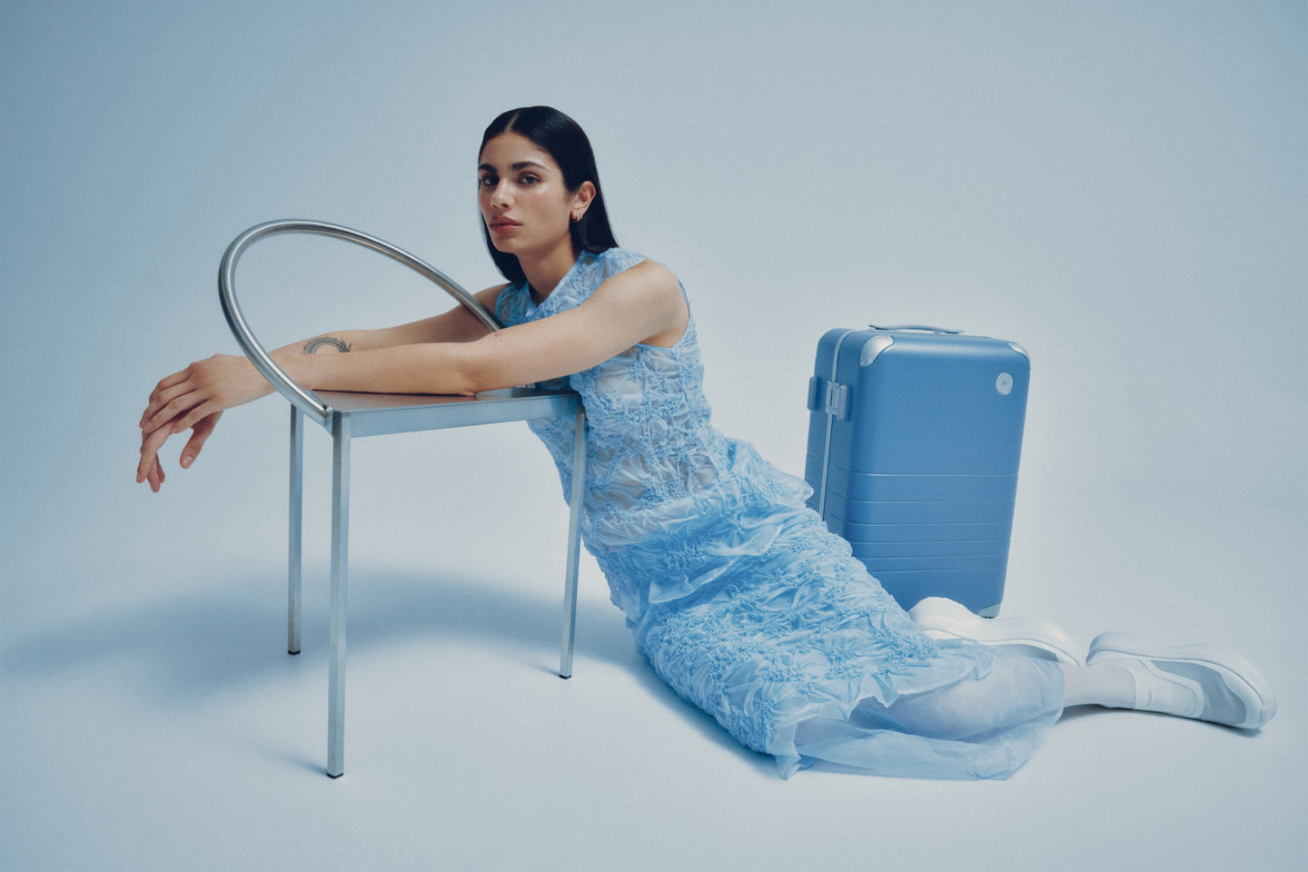 A model in a blue lace dress leans on a minimalist metal chair, with a Hybrid Carry-On in Elemental Blue beside her.