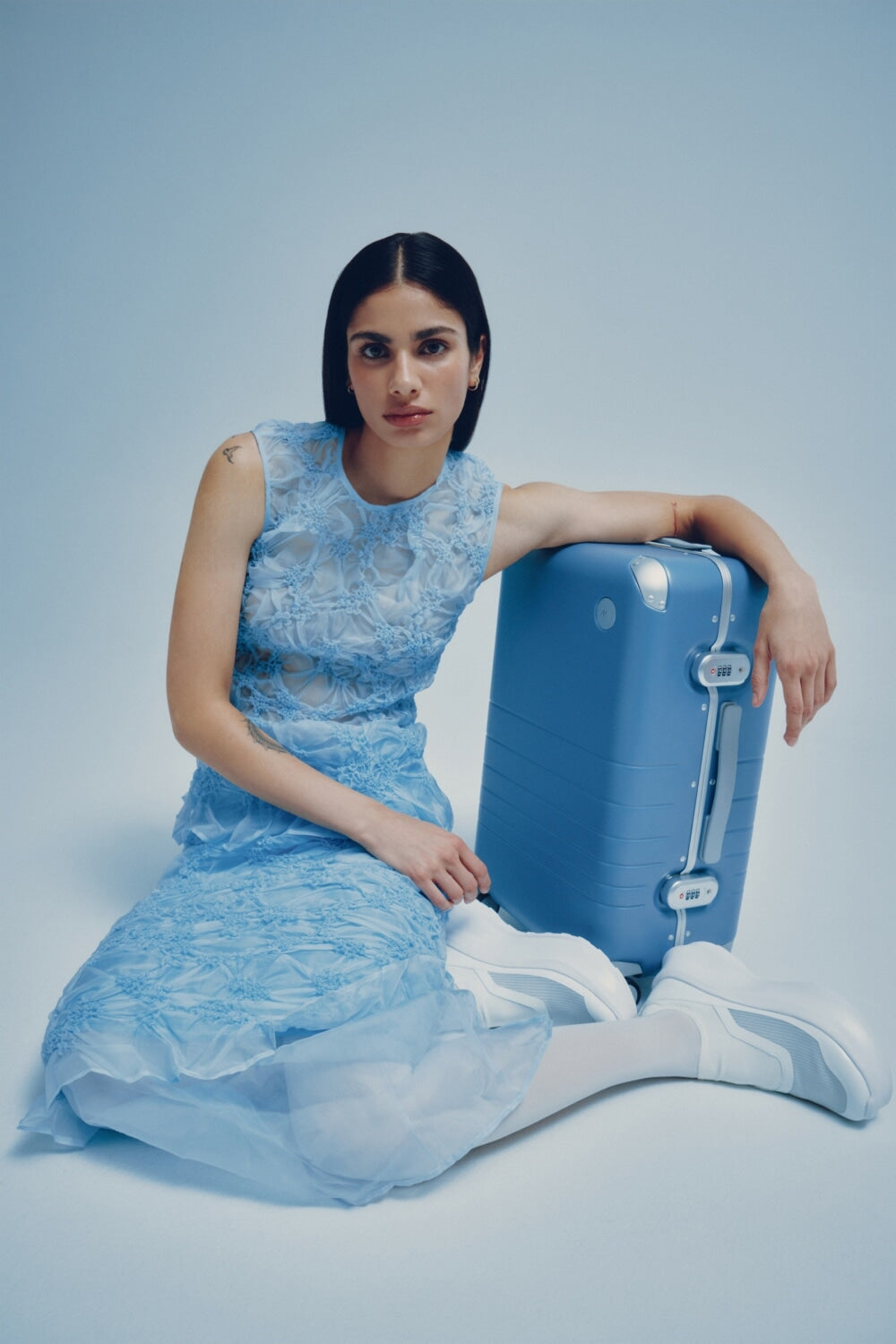 A model in a blue lace dress sits on the floor, resting her arm on a Hybrid Carry-On in Elemental Blue, looking directly at the camera.