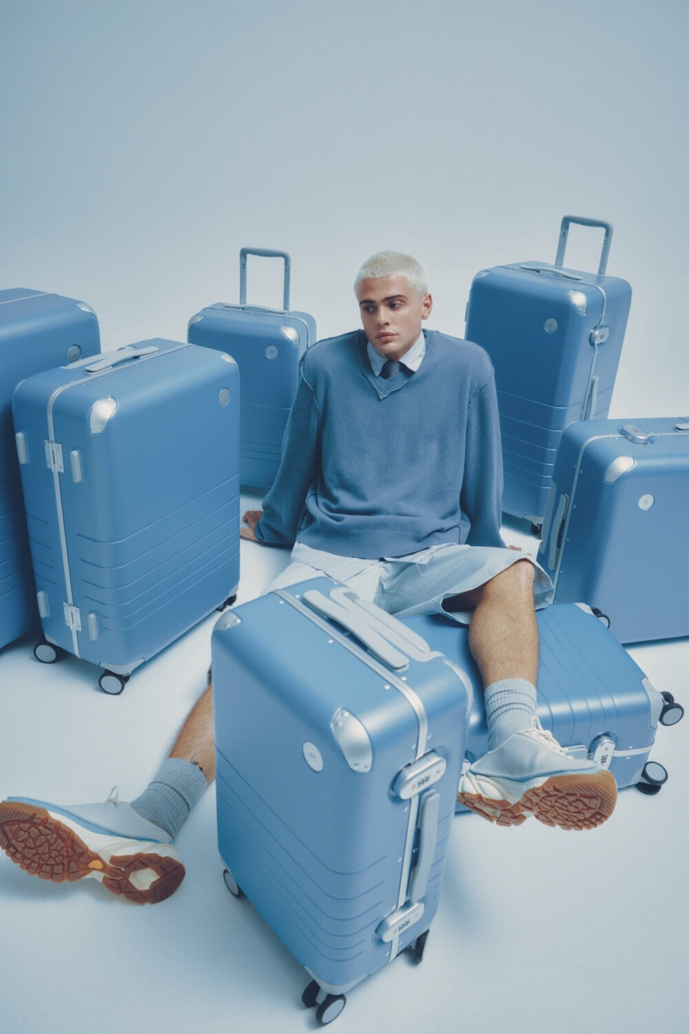 A model in a blue outfit sits surrounded by Hybrid luggage in Elemental Blue, resting one leg on a suitcase, against a soft blue backdrop.