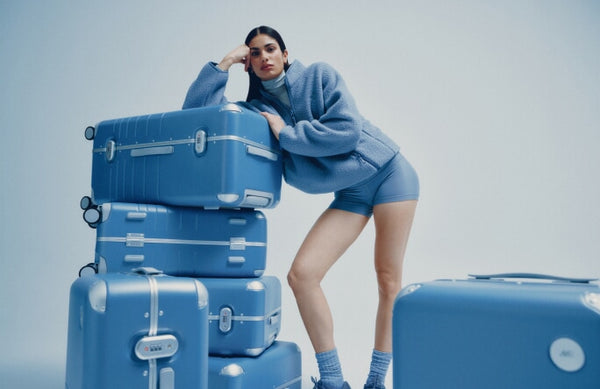 A model in a blue fleece jacket and shorts leans on a stack of Hybrid suitcases in Elemental Blue, gazing at the camera.