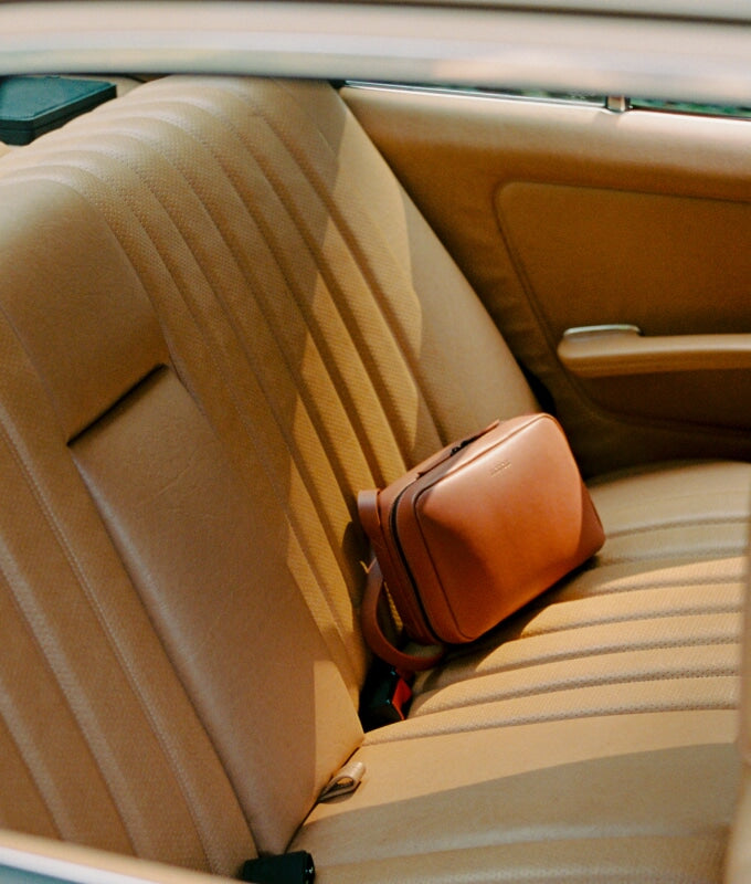 A metro crossbody in mahogany (vegan leather) placed on the backseat of a vintage car with beige upholstery.