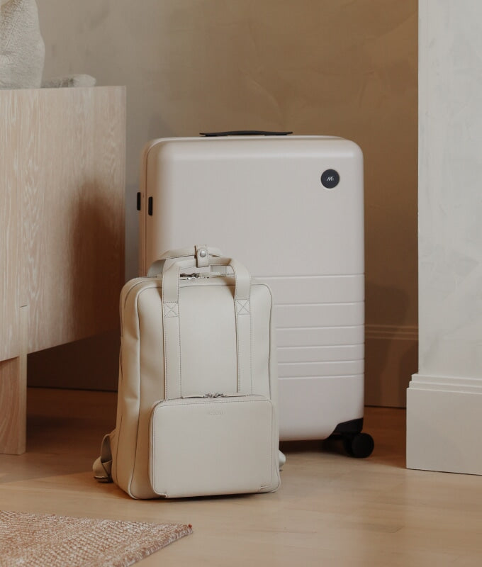 A check-in medium in desert taupe and metro backpack in ivory (vegan leather) placed on a light wooden floor in a minimalist home interior.