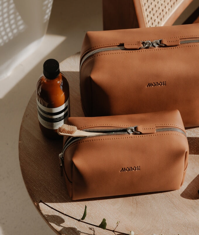 A small and large metro toiletry case in mahogany (vegan leather) embossed with the Monos logo sitting on a wooden surface alongside a bottle of skincare product and a toothbrush, bathed in warm natural light.