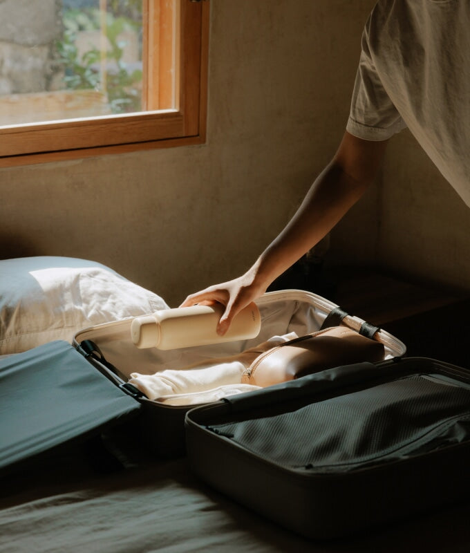 A hand carefully packing a kiyo uvc water bottle in castle rock into an open carry-on on a bed, with soft natural light streaming in through a wooden-framed window.