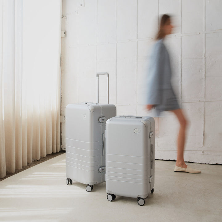 A woman in motion walks past the Hybrid Trunk Medium and Trunk suitcases in Silver, standing in a softly lit, minimalist room.