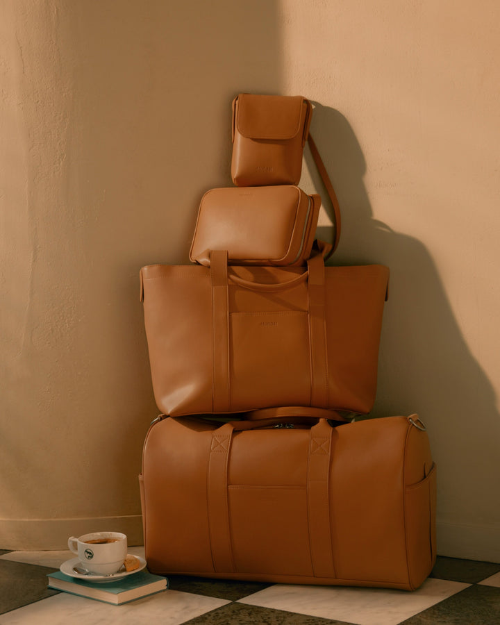 A neatly stacked set of Monos bags, all in Saddle Tan (Vegan Leather), sits against a soft, tan wall.  The stack includes a Metro Mini Crossbody at the top, Metro Crossbody in the middle, and a Metro Tote and Metro Carry-All Duffel at the bottom. A cup of coffee rests beside the bags on a book, completing the cozy scene.