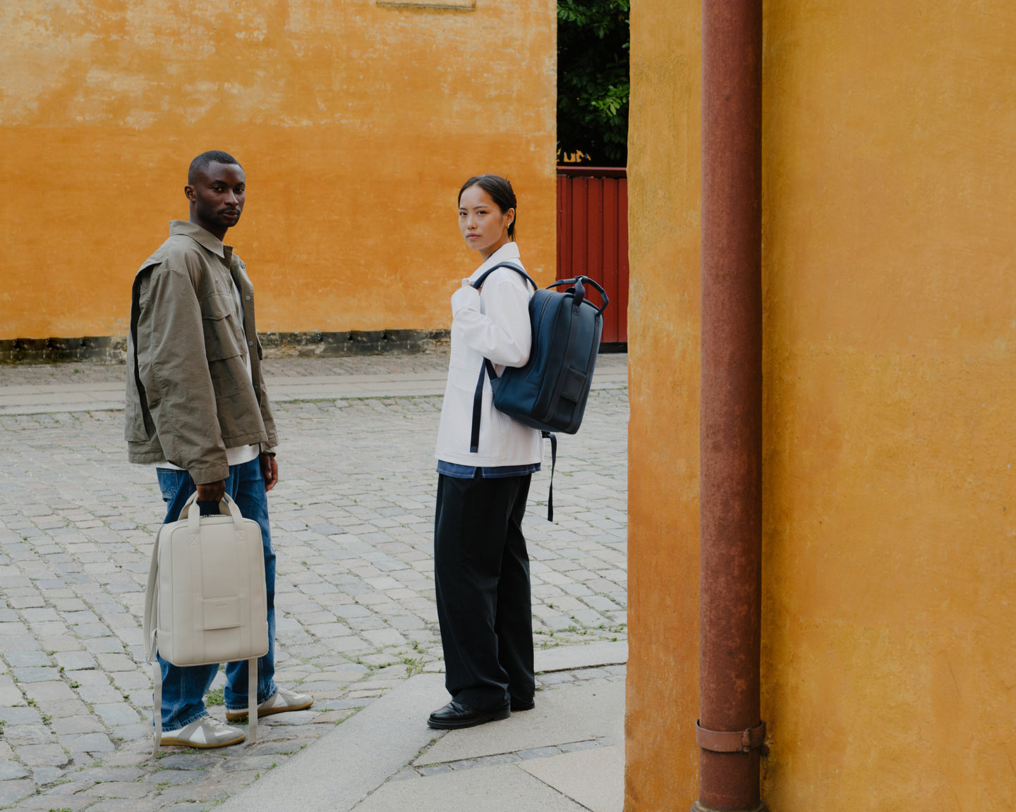 Two individuals standing in a cobblestone courtyard, one holding a Metro Backpack in Ivory (Vegan Leather) and the other wearing a Metro Backpack in Oxford Blue. The Metro Classic Kit is not attached to the front of either backpack. The backdrop features a rustic orange wall.