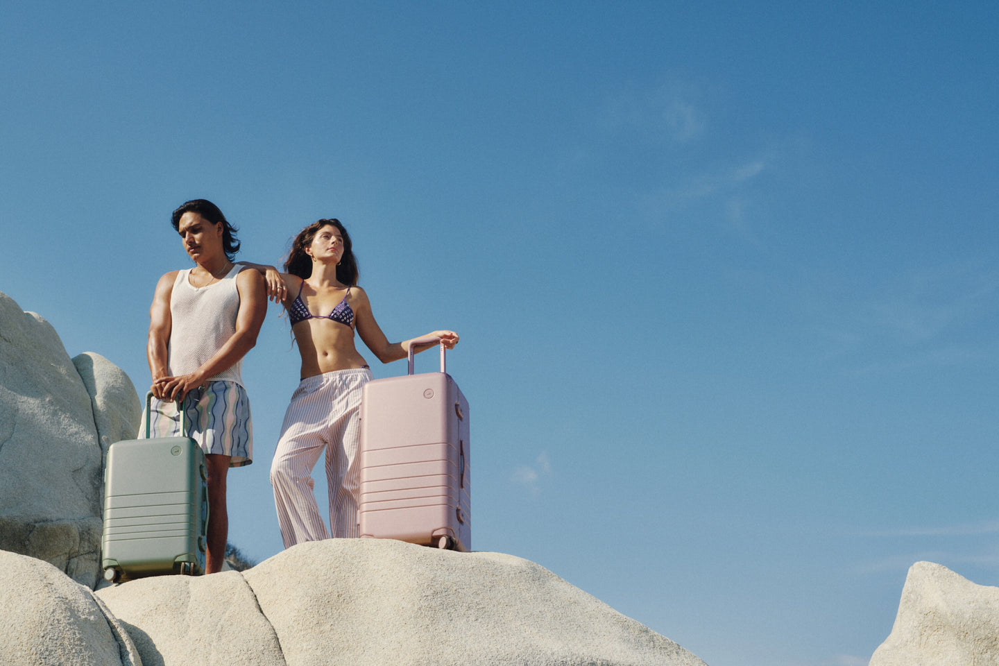 Two travelers stand on sunlit rocks with the Hybrid Carry-On in Mint and the Hybrid Trunk in Orchid, basking in the calm of a clear blue sky.