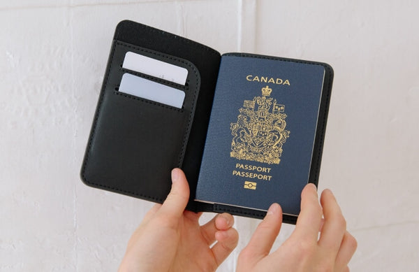 A black leather passport holder with card slots, shown open with a Canadian passport inside, held by two hands against a white textured wall.