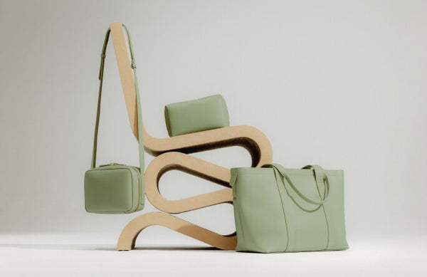 A minimalist wooden chair is styled with Metro Tote, Metro Crossbody, and Large Metro Toiletry Case in Cactus.