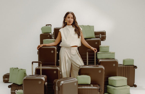 A woman stands among stacked Hybrid suitcases in Adobe Brown and bags and accessories in Cactus