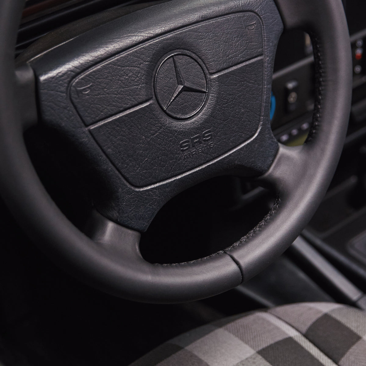 Close-up of a black leather Mercedes-Benz steering wheel with the brand's three-pointed star emblem in the center, set inside a car cabin with a gray plaid seat visible in the lower right.