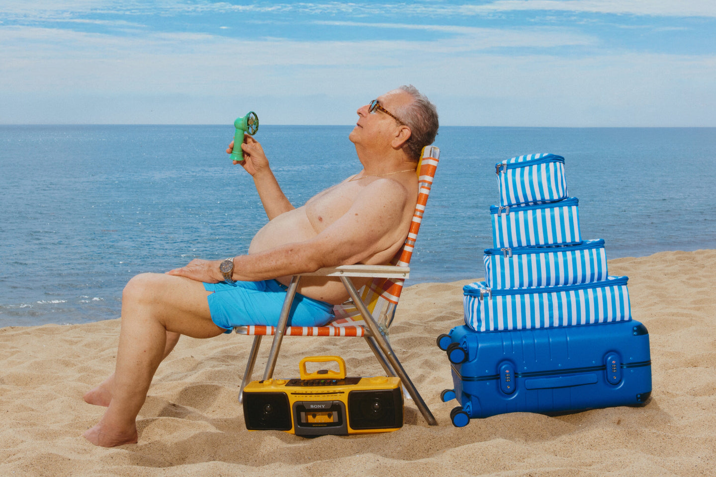 Man sitting on a beach chair with a blue suitcase and stack of packing cubes as well as a boombox, holding up a portable fan.