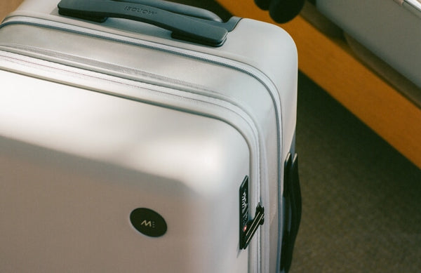 A close-up of an expandable carry-on in storm grey featuring a TSA-accepted lock, a Monos logo, placed indoors with warm lighting.