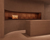  The Metro Tote in Saddle Tan displayed within a recessed wood alcove inside the Monos SoHo store, framed by curved terracotta brick walls and warm ambient lighting.