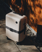 Monos Carry-On in Desert Taupe with Luggage Strap in Midnight Black under soft sunlight.