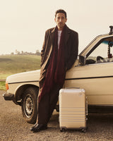 Adrien Brody stands beside a vintage cream-coloured car on a rural road, with a Monos aluminum carry-on plus in aspen silver by his feet and rolling green hills in the background.