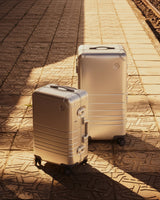 Two Monos aluminum suitcases in Aspen Silver stand on a textured train platform, partially illuminated by sunlight, with railway tracks visible in the background.