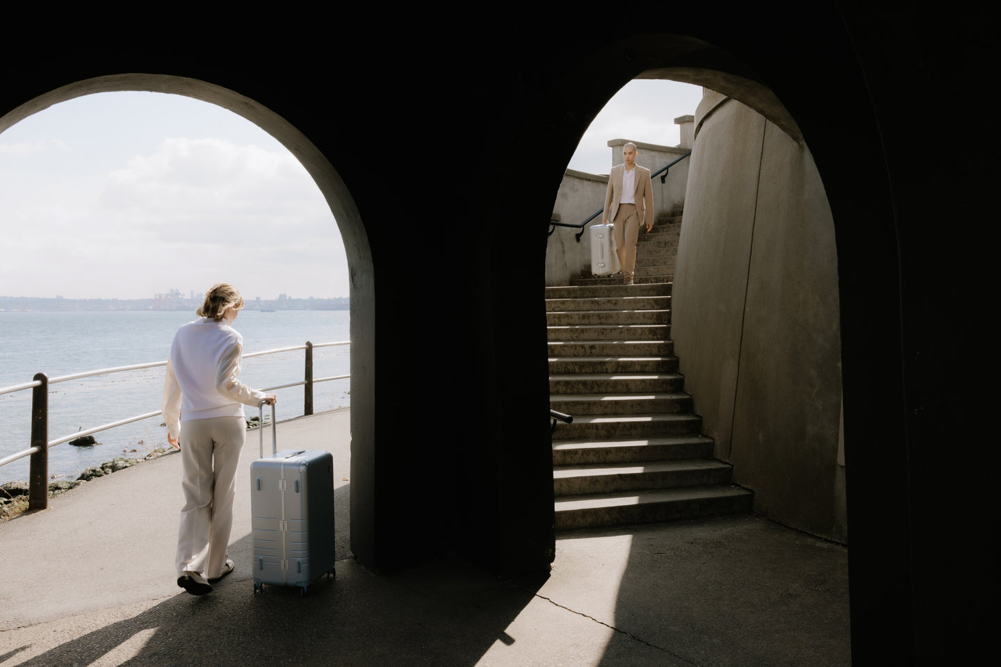 A woman in white walks along a waterfront path with a hybrid trunk in silver, while a man in a beige suit descends stairs carrying a hybrid carry-on in silver, framed by dark archways.