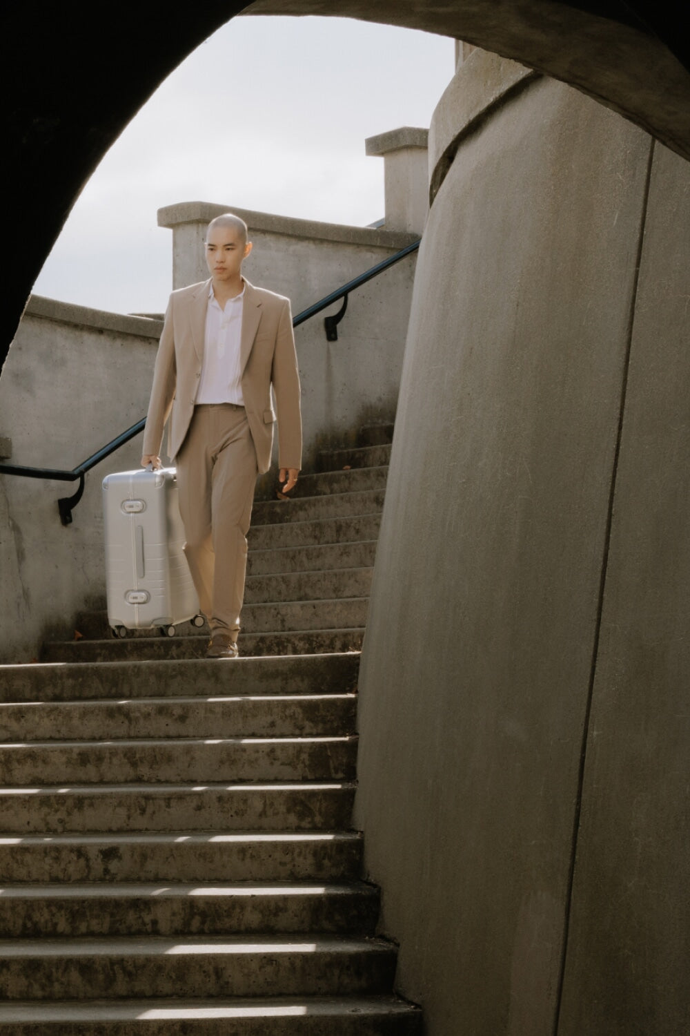 A man in a beige suit descends stairs carrying a hybrid carry-on in silver, framed by dark archways.