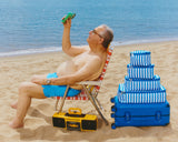 Man sitting on a beach chair with a blue suitcase and stack of packing cubes as well as a boombox, holding up a portable fan.