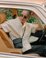 A man wearing sunglasses and a relaxed cream-coloured shirt sitting in the driver's seat of a vintage car, wearing a metro mini crossbody in carbon black (vegan leather)