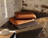 Two metro laptop sleeves in mahogany (vegan leather) embossed with the Monos logo, neatly stacked on a rustic wooden desk, with a sleek laptop partially tucked inside, illuminated by soft natural light.