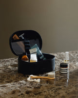 A metro cosmetic case in carbon black (vegan leather) opened to reveal neatly organized personal care items, sitting on a marble surface alongside a toothbrush, skincare bottles, and grooming essentials.