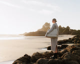 A woman dressed in a long, light-grey sweater dress and black boots standing on rocky terrain at a serene beach during golden hour, carrying a Metro Weekender in Ivory (Vegan leather).