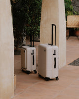 A carry-on pro and check-in medium in desert taupe standing side by side on a tiled patio surrounded by lush greenery.