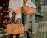 Two individuals standing in a vintage-style shop holding matching Saddle Tan (vegan leather) bags, one a metro carry-all duffel and the other a metro tote