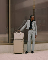 A woman dressed in a tailored grey suit standing in an urban setting with a hybrid carry-on in champagne and a matching metro tote in ivory (vegan leather) perched on top.