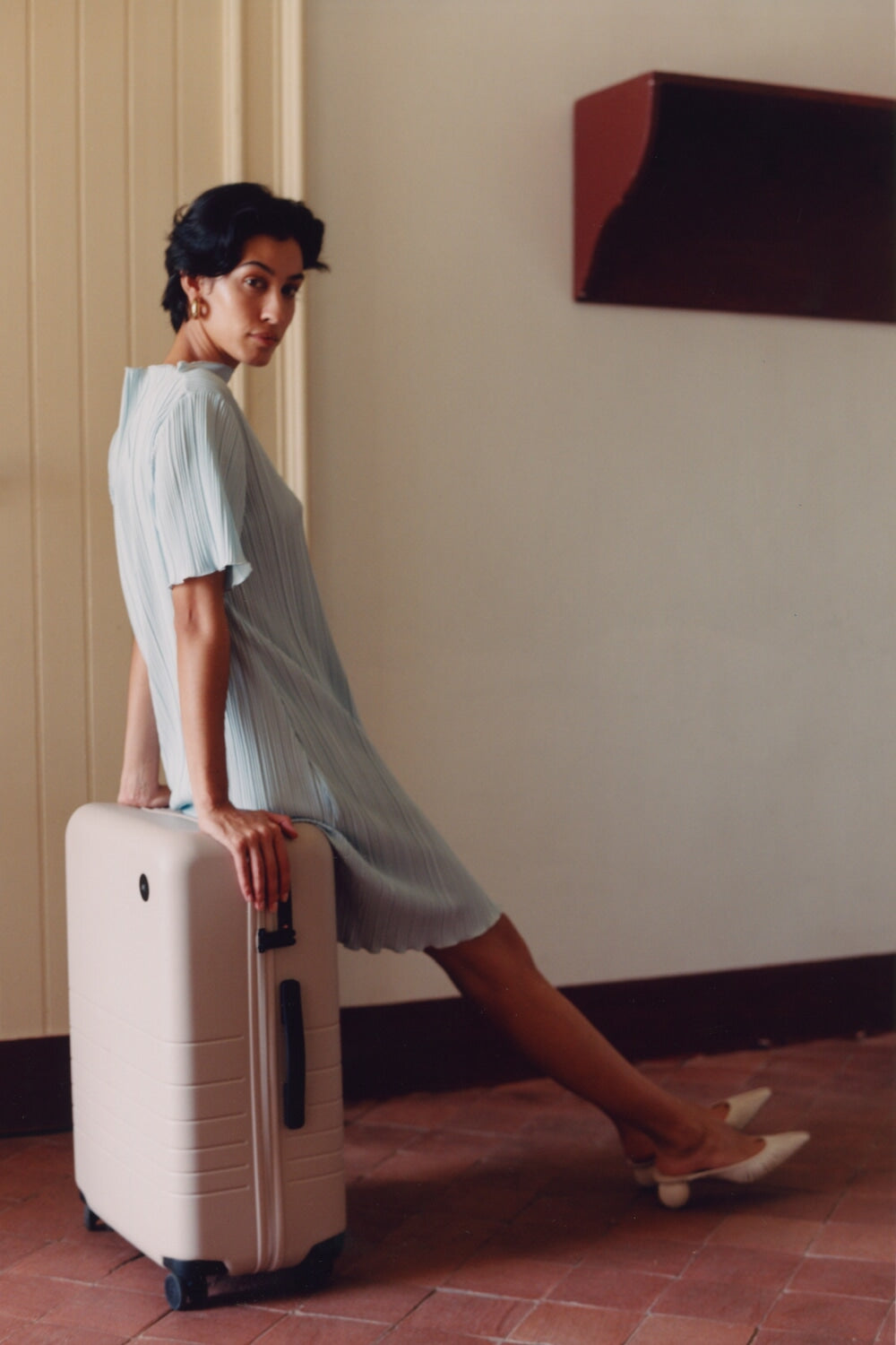 A woman in a light blue pleated dress sitting on a Check-In Medium in Desert Taupe. She looks back over her shoulder, adding a touch of elegance and sophistication.