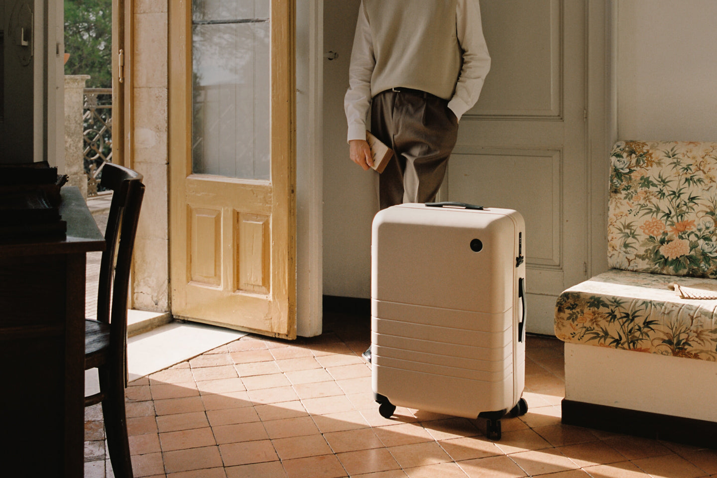 A check-in large in desert taupe placed near an open doorway in a cozy, vintage-style room. A person stands nearby, dressed in a neutral outfit, holding a small book.